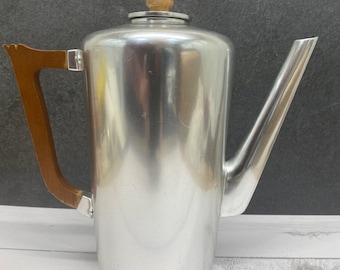 Puede incluir: Una cafetera de aluminio plateado con un mango y un pomo de tapa de madera. La cafetera tiene un diseño elegante y moderno.