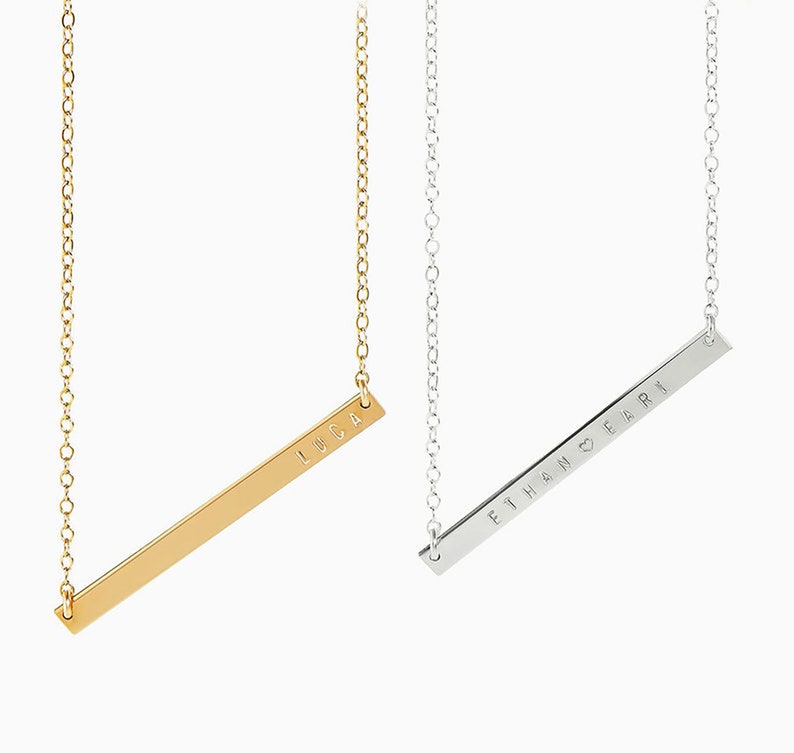 May include: Two gold and silver bar necklaces with personalized names. The gold necklace has the name "LUCA" engraved on the bar. The silver necklace has the name "ETHAN HEART EARI" engraved on the bar.