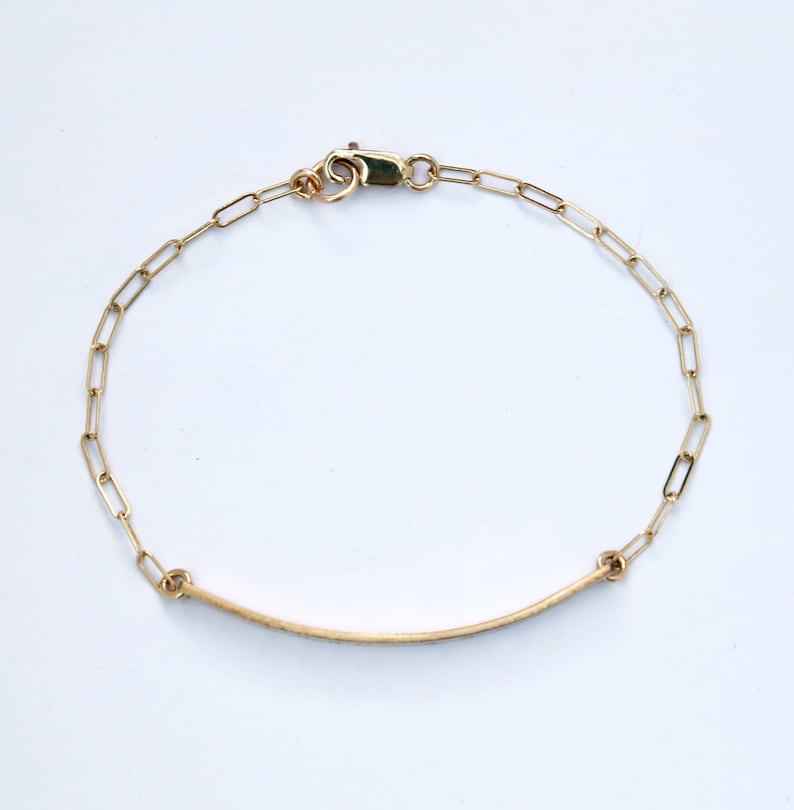 Puede incluir: Un brazalete de cadena de oro con un colgante de barra curva delgado.