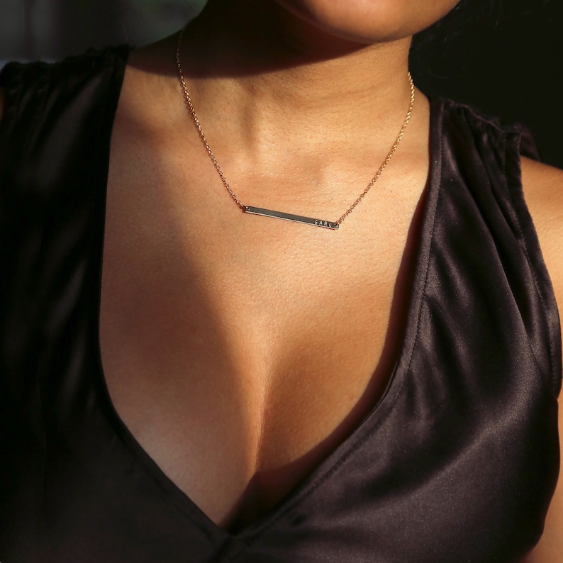 May include: A gold chain necklace with a thin, horizontal bar pendant. The bar is engraved with the word "LOVE". The necklace is worn by a person with a low-cut neckline.