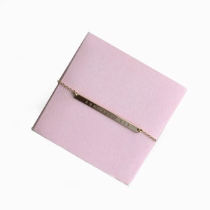 May include: A gold bar necklace with the name "DOMINIC & LUCA" engraved on it. The necklace is displayed on a pink box.