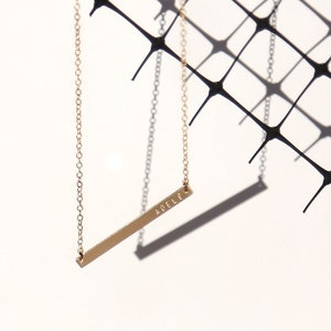 May include: A gold chain necklace with a thin, horizontal bar pendant engraved with the name "AD&Egrave;LE".