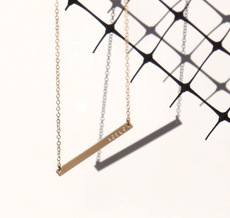 May include: A gold chain necklace with a thin, horizontal bar pendant engraved with the name "AD&Egrave;LE".
