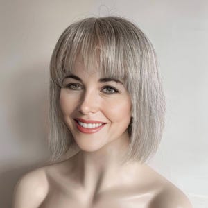 May include: A smiling person with light skin and gray hair styled in a short bob with bangs. The person is wearing makeup, including lipstick. The background is a neutral color.