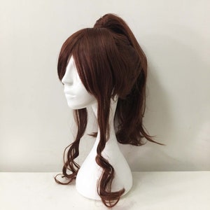 K&ouml;nnte beinhalten: Eine braune Per&uuml;cke mit langen, lockigen Haaren, die zu einem hohen Pferdeschwanz gebunden sind. Die Per&uuml;cke befindet sich auf einem wei&szlig;en Mannequin-Kopf.