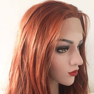 May include: A mannequin head with a long, layered, auburn wig. The wig features a natural-looking hairline and is styled with loose waves. The mannequin has full eyelashes and red lipstick, with a neutral background.