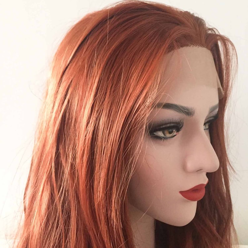 May include: A mannequin head with a long, layered, auburn wig. The wig features a natural-looking hairline and is styled with loose waves. The mannequin has full eyelashes and red lipstick, with a neutral background.