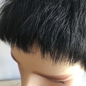 May include: Close-up of a person's black hair, styled in a short, straight cut. The hair is parted on the side and falls just above the forehead.
