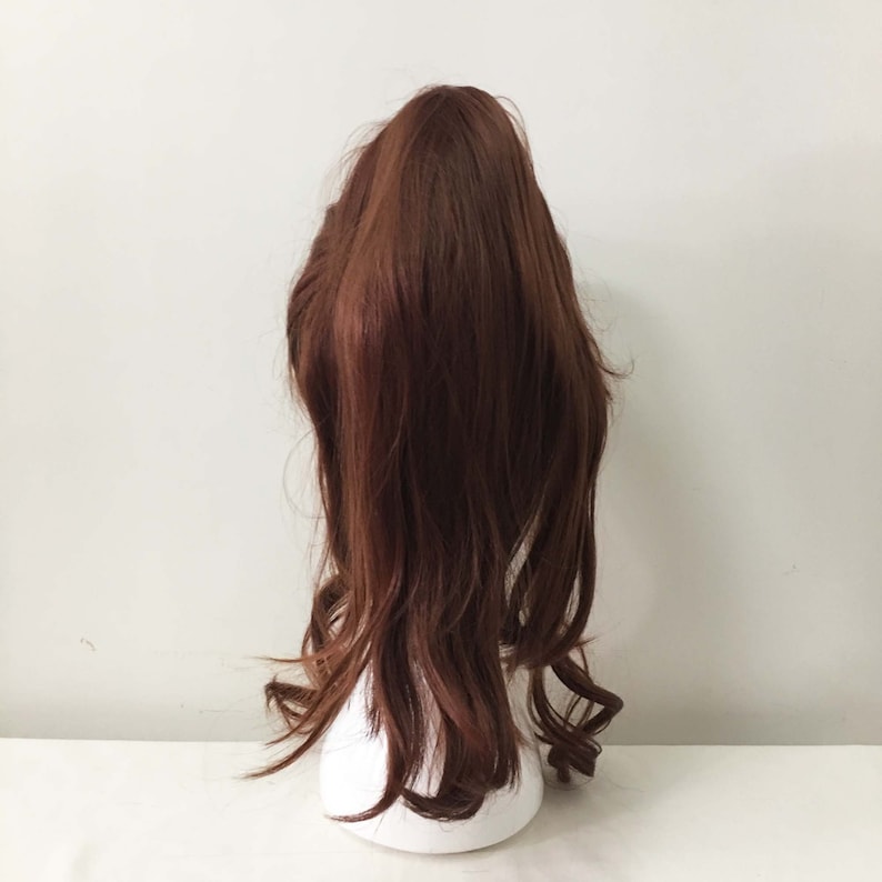 K&ouml;nnte beinhalten: Eine lange, braune, gewellte Pferdeschwanz-Haarverl&auml;ngerung. Das Haar ist locker und l&auml;ssig gestylt, mit einigen Str&auml;hnen, die um das Gesicht fallen.