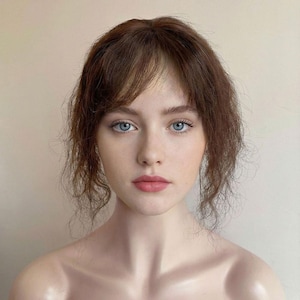 May include: A portrait of a person with fair skin, light blue eyes, and reddish-brown hair styled with bangs. The person has a neutral expression and is looking directly at the viewer. The background is a solid, light beige color.