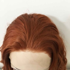 May include: A close-up of a light brown wig with a lace front. The wig is styled in loose waves.