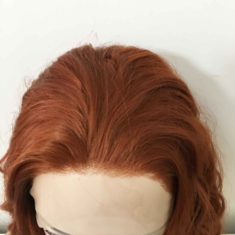 May include: A close-up of a light brown wig with a lace front. The wig is styled in loose waves.