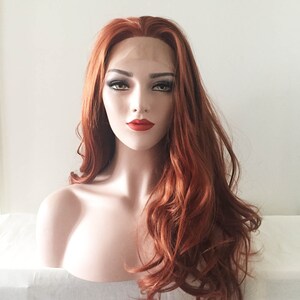 May include: A mannequin head displaying a long, wavy auburn wig. The wig features a lace front and a natural-looking hairline. The mannequin has a fair complexion, red lipstick, and dark eye makeup. The background is a plain white.