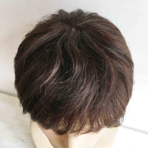 May include: A close-up of a dark brown wig with a short, layered style. The wig is styled with a side part and has a natural, realistic look.