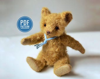 Op de afbeelding: Een kleine, handgemaakte teddybeer met lichtbruine vacht, een blauw lint om de nek en donkere ogen. De beer zit met uitgestrekte armen en benen. Een blauwe cirkel met de tekst "PDF PATTERN" staat linksboven.