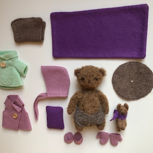 Puede incluir: Un oso de peluche marrón que lleva un chaleco gris y zapatillas moradas. El oso está rodeado de una variedad de prendas de fieltro, incluyendo una chaqueta verde, un chaleco morado, un gorro rosa y una manta morada.