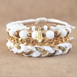 Op de afbeelding: Een gelaagde armband met witte en gouden kralen, een gouden ketting en een gevlochten wit en gouden koord. De armband heeft een gouden hamsa-handbedel.