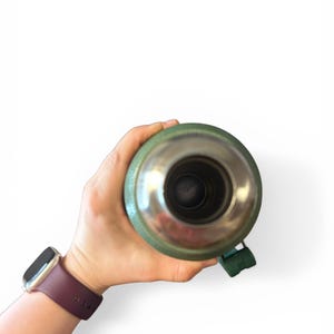 May include: A green and silver insulated flask held in a hand. The flask has a silver interior and a green exterior with a green handle. A maroon watch is on the wrist.