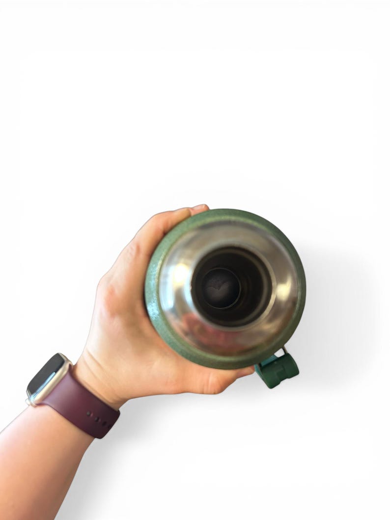 May include: A green and silver insulated flask held in a hand. The flask has a silver interior and a green exterior with a green handle. A maroon watch is on the wrist.