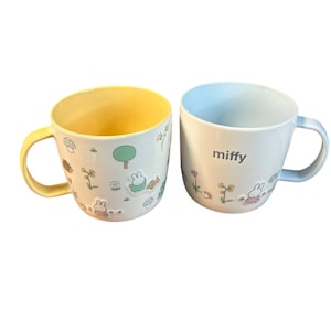 May include: Two white ceramic mugs with colorful interiors and handles. One mug has a yellow interior and handle, the other has a light blue interior. Both mugs feature a cartoon bunny and floral design, with the word "miffy" on one.