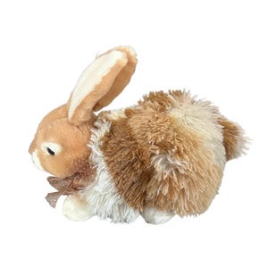 Puede incluir: Un peluche de conejo en tonos marrón, beige y blanco. El conejo tiene orejas largas, un cuerpo esponjoso y un pequeño lazo alrededor del cuello. El juguete está diseñado para parecerse a un conejo real.