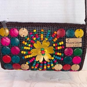 May include: A rectangular brown purse with a woven texture, adorned with colorful circular accents and a central flower design. The purse features a shoulder strap and a small wooden tag that reads "Punta Cana".