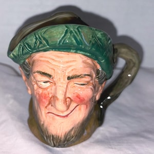 May include: A ceramic mug shaped like a smiling man's face, with a green hat and a brown handle. The mug has a detailed, hand-painted design with a rosy complexion and a gray beard. The mug is likely vintage or antique.