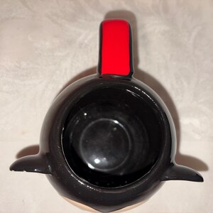 May include: A black ceramic mug shaped like a penguin, with a red rectangular accent on top. The mug has a glossy finish and a small, rounded base. The interior is black, and the penguin's belly is a light beige color.