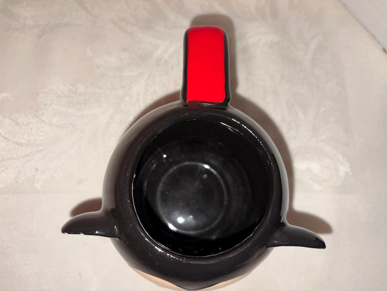 May include: A black ceramic mug shaped like a penguin, with a red rectangular accent on top. The mug has a glossy finish and a small, rounded base. The interior is black, and the penguin's belly is a light beige color.