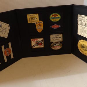 May include: A black display case with various vintage food labels. Labels include "Maple Hill Vermont Syrup", "Pasier's", "IDYL Tomato Catsup", and "Smile". The case is open, revealing multiple compartments.