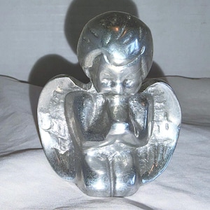 May include: A silver angel figurine with folded hands and detailed wings. The angel is in a praying position, with a serene expression. The sculpture is made of a shiny, metallic material, likely a decorative item.