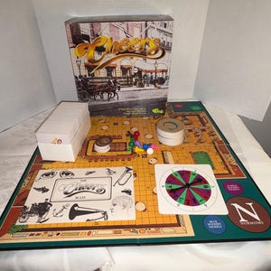 May include: A vintage Cheers board game with a box featuring a street scene and the game's logo. The game board is yellow and green, with various game pieces, cards, and a spinner. A white box of cards is also present.
