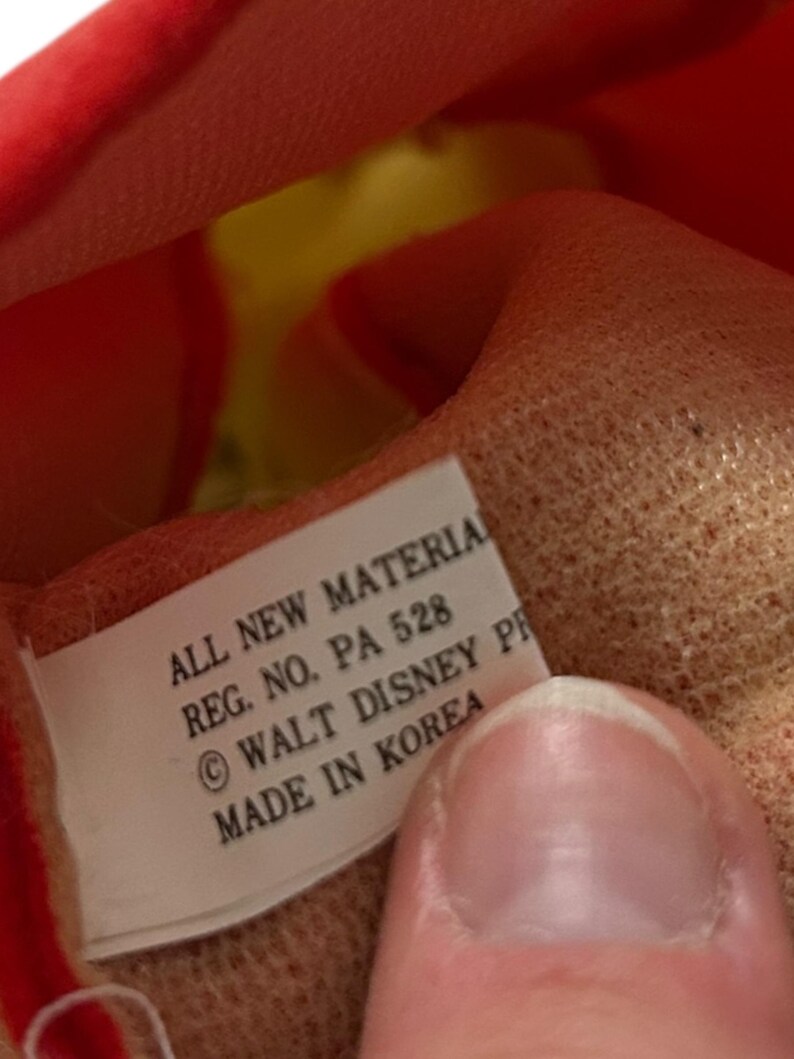 以下が含まれることがあります： 「ALL NEW MATERIAL, REG. NO. PA 528, &copy; WALT DISNEY, MADE IN KOREA」と書かれたタグのクローズアップ。赤い布と黄色の布で作られたぬいぐるみです。
