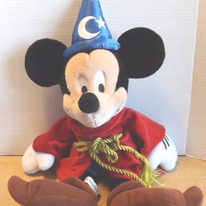 May include: A plush Mickey Mouse dressed as a sorcerer, wearing a blue hat with a crescent moon and stars. He has a red robe with a gold rope belt and brown shoes. The plush toy has black ears, a white face, and white gloves.