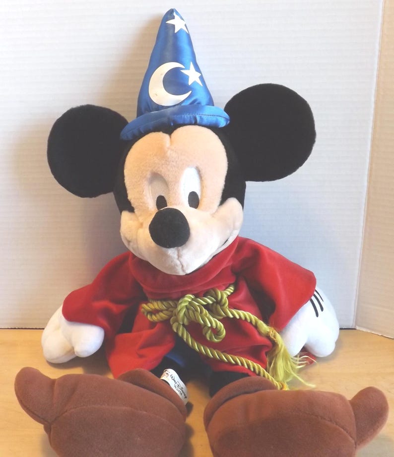 May include: A plush Mickey Mouse dressed as a sorcerer, wearing a blue hat with a crescent moon and stars. He has a red robe with a gold rope belt and brown shoes. The plush toy has black ears, a white face, and white gloves.