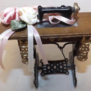 May include: A detailed miniature sewing machine. The machine has a black body, gold accents, and a wooden table. Pink and white ribbons and a small green shoe are attached to the table. The base is black metal.