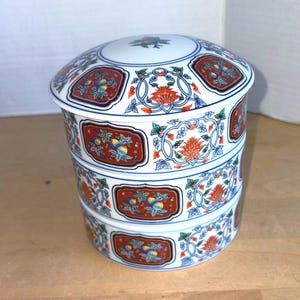 May include: A three-tiered, white porcelain container with a lid. The container is adorned with intricate floral and fruit designs in red, orange, blue, and green. The design is reminiscent of traditional Asian art. The container is likely used for food storage or display.
