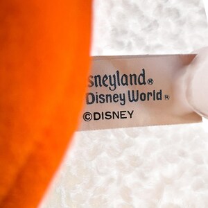 以下が含まれることがあります： 「Disneyland」と「Disney World」の文字とディズニーのロゴが入ったタグのクローズアップ。タグは白いテクスチャードファブリックに付いています。画像の左側にはオレンジ色の物体が見えます。