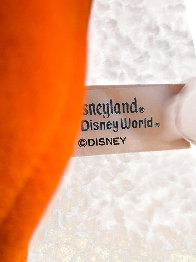 以下が含まれることがあります： 「Disneyland」と「Disney World」の文字とディズニーのロゴが入ったタグのクローズアップ。タグは白いテクスチャードファブリックに付いています。画像の左側にはオレンジ色の物体が見えます。