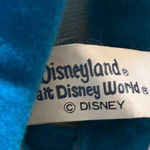 以下が含まれることがあります： 「Disneyland」と「Walt Disney World」の文字が濃い青色で印刷されたタグのクローズアップ。タグには著作権マークと「DISNEY」の文字も含まれています。タグは明るい青色の生地で部分的に隠されています。
