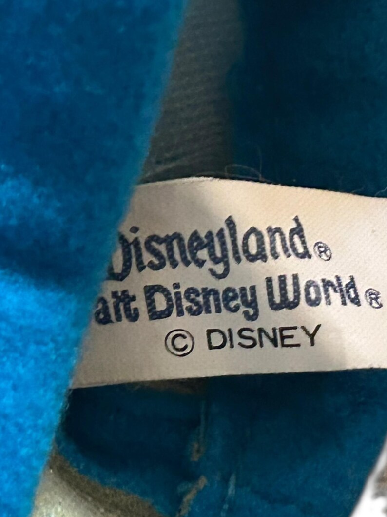 以下が含まれることがあります： 「Disneyland」と「Walt Disney World」の文字が濃い青色で印刷されたタグのクローズアップ。タグには著作権マークと「DISNEY」の文字も含まれています。タグは明るい青色の生地で部分的に隠されています。
