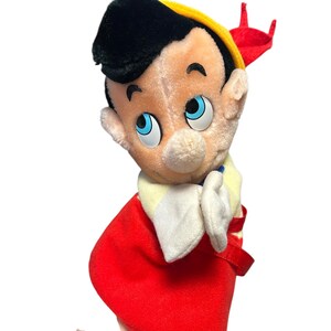 以下が含まれることがあります： 黒い髪、黄色の帽子、赤いアクセントのぬいぐるみピノキオ。 青い目、ベージュの顔、白い手袋があります。赤い衣装が見えます。
