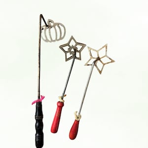 May include: Three vintage metal rosette irons with wooden handles. One iron has a pumpkin shape, and the other two have star shapes. The handles are red and black, with twine and pink ribbon accents.