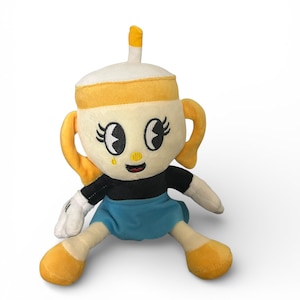 May include: A plush toy character with a cup-like head, yellow handle-like ears, and a small yellow straw. The character has a black shirt, blue skirt, and yellow feet. The face has large eyes and a small smile. The toy is on a white background.
