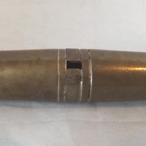 Puede incluir: Un silbato antiguo de latón. El cuerpo cilíndrico tiene una ligera forma de reloj de arena, con una banda central con líneas grabadas y una abertura rectangular. El latón presenta una pátina cálida y envejecida.