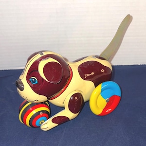 May include: Vintage wind-up toy dog with a cream-colored body, burgundy spots, and a long tail. The dog has blue eyes and a red collar. It has two multi-colored wheels and a striped ball. A classic collectible toy.