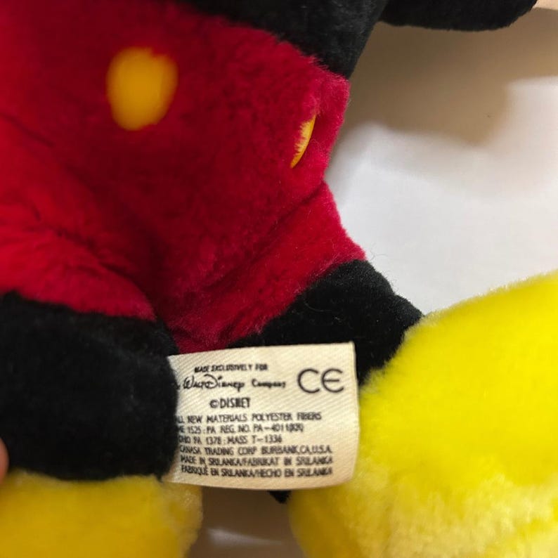 Puede incluir: Primer plano de un peluche de Mickey Mouse. El juguete tiene un cuerpo rojo, brazos y piernas negros y guantes y pies amarillos. Una etiqueta con texto es visible.