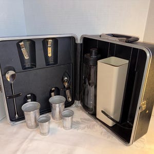 May include: An open vintage cocktail set in a black case with silver trim. The case contains two compartments with bar tools, including a shaker, jiggers, and shot glasses. The set is designed for mixing drinks.