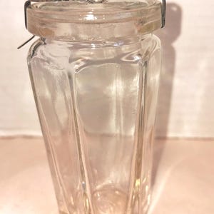 Pode incluir: Um frasco de vidro transparente e vintage com um corpo facetado e uma tampa de metal articulada. O frasco tem uma forma retangular com laterais anguladas e uma base hexagonal. A tampa é fixada com um fecho de arame.