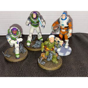 Puede incluir: Cuatro figuras de acción de plástico de la película Lightyear. Dos figuras llevan trajes espaciales blancos y verdes, una lleva un traje de vuelo naranja y blanco con casco, y una lleva un traje táctico verde con un arma y bolsas marrones.